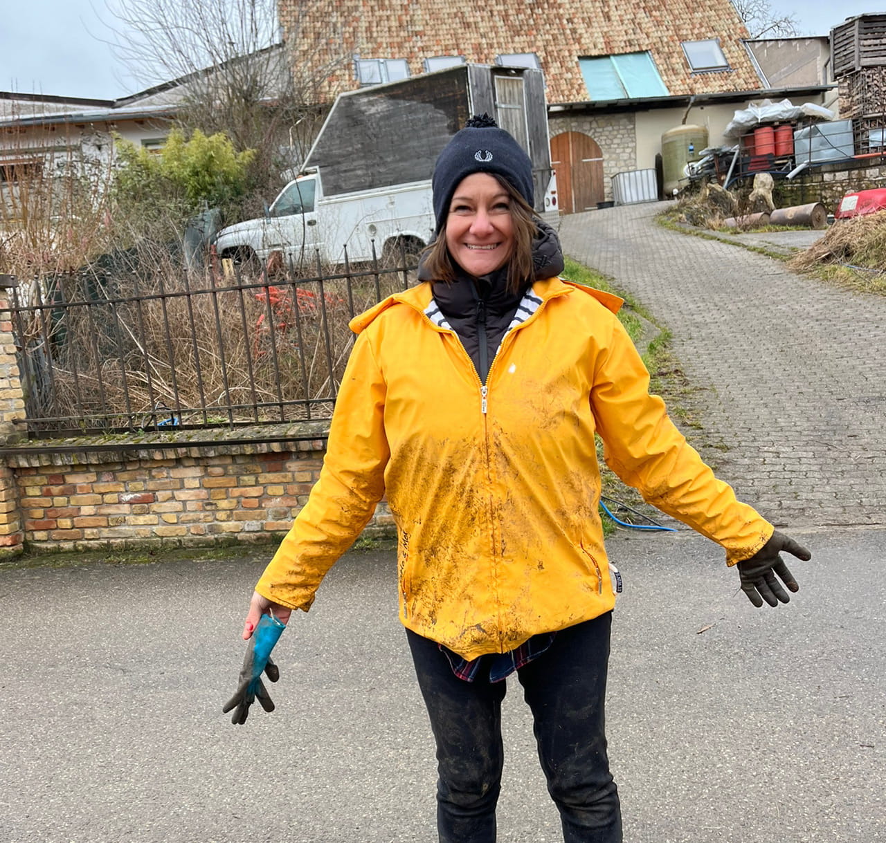 Authentisches Porträt von Claudia diCarlo, der Besitzerin des Radlertreffs, die mit weit geöffneten Armen auf einer asphaltierten Dorfstraße steht. Sie trägt eine auffällige, mit Matsch bespritzte gelbe Regenjacke, schwarze Matschhosen, eine schwarze Beanie-Mütze mit Logo und schwarze Handschuhe. In ihrer linken Hand hält sie einen zusätzlichen blauen Arbeitshandschuh. Sie strahlt und blickt direkt in die Kamera.